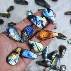 May include: A collection of labradorite gemstone coffin pendants in various colors, including blue, gold, and green. The pendants are faceted and have a polished finish. Some pendants have a hole drilled at the top for stringing.
