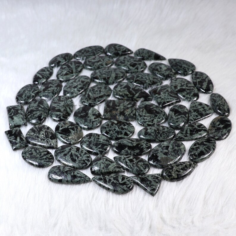 May include: A collection of polished gemstones in various shapes, including ovals, teardrops, and rectangles. The stones are a dark green color with black veining, arranged on a white, fluffy surface.