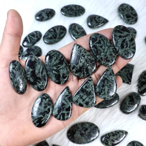 May include: A collection of polished, dark green gemstones with black veining. The stones are in various shapes, including ovals, teardrops, and irregular forms. The gemstones are displayed on a white, fluffy surface.
