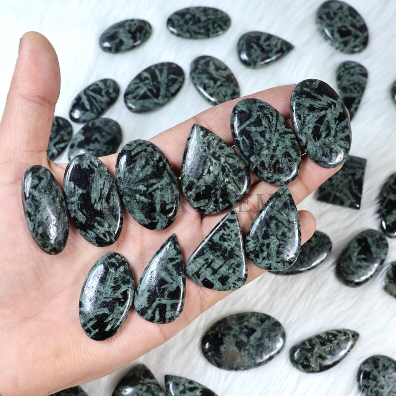 May include: A collection of polished gemstones in various shapes, including ovals, teardrops, and rectangles. The stones are a dark green color with black veining, creating a unique pattern. The gemstones are displayed on a white surface.