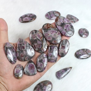 May include: A collection of polished gemstones in various shapes, including oval, teardrop, and rectangular. The stones feature a mix of purple, gray, and brown colors with a speckled pattern. The image includes the text "NIKHAT GEMS".