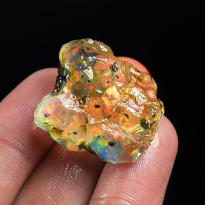 Può includere: Una gemma di opale grezza con una vibrante gamma di colori. La pietra presenta una miscela di tonalità arancioni, gialle, verdi e blu, con inclusioni scure. L'opale è tenuto in una mano, mostrando il suo stato naturale e non lucidato.