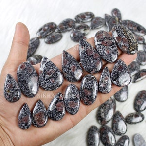 Peut inclure: Une collection de pierres précieuses polies de différentes formes, dont des gouttes et des ovales. Les pierres présentent un motif gris et noir moucheté avec des inclusions rouges, présentées sur une surface blanche.