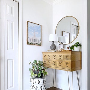 May include: A wooden cabinet with 12 drawers on hairpin legs, a round gold mirror, a white lamp with a gray ceramic base, a potted plant, and a white and black patterned stool. The cabinet is in a corner of a room with white walls and hardwood floors.