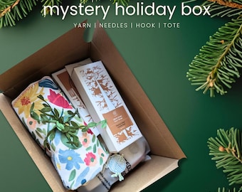 May include: A brown cardboard box labeled "mystery holiday box" containing yarn, needles, a hook, and a tote. The box includes a floral fabric item, two white boxes with brown tree designs, and a yarn-themed tag. The background is a dark green with pine branches.