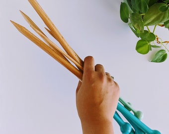 May include: A set of four wooden knitting needles with turquoise colored tips. The needles are held in a hand.