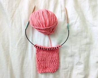 May include: A pink yarn ball with knitting needles and a knitted swatch. The needles are wooden with black cables and teal connectors. The knitted swatch is a solid pink color, showing the texture of the knit stitches.
