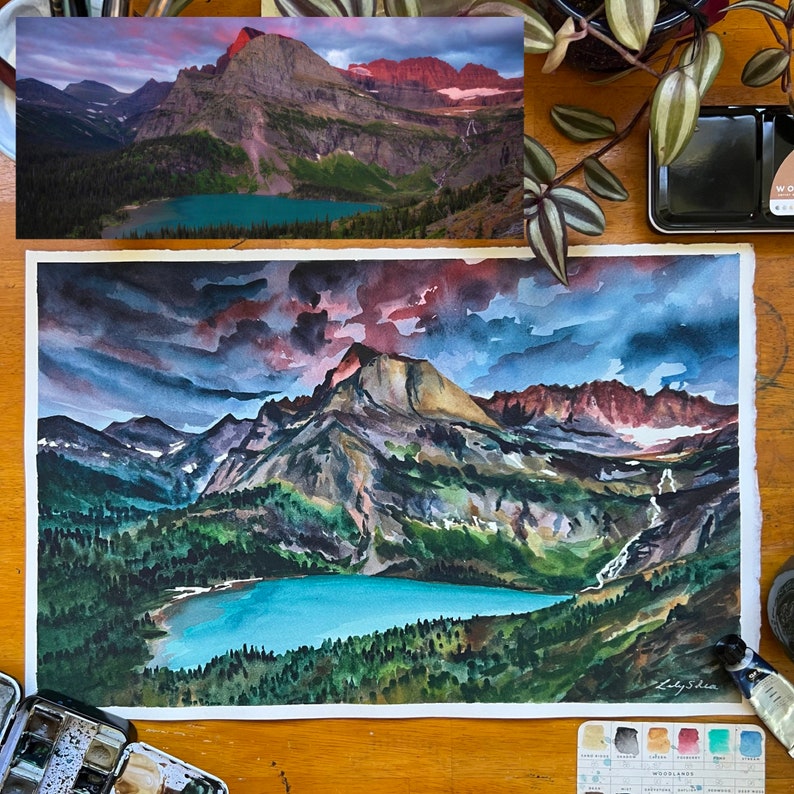 Puede incluir: Pintura acuarela de una cordillera con un lago en primer plano. El cielo es de un azul dram&aacute;tico con nubes oscuras y un toque de rosa en la distancia. Las monta&ntilde;as son una mezcla de verde y marr&oacute;n, con algunos picos rojos. El lago es de un azul turquesa brillante.