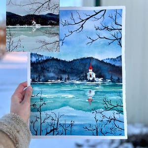 Puede incluir: Pintura acuarela de una iglesia con techo rojo en un lago con monta&ntilde;as cubiertas de nieve en el fondo. La pintura est&aacute; enmarcada y sostenida por una mano.