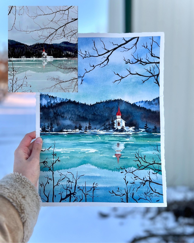 Puede incluir: Pintura acuarela de una iglesia con techo rojo en un lago con monta&ntilde;as cubiertas de nieve en el fondo. La pintura est&aacute; enmarcada y sostenida por una mano.