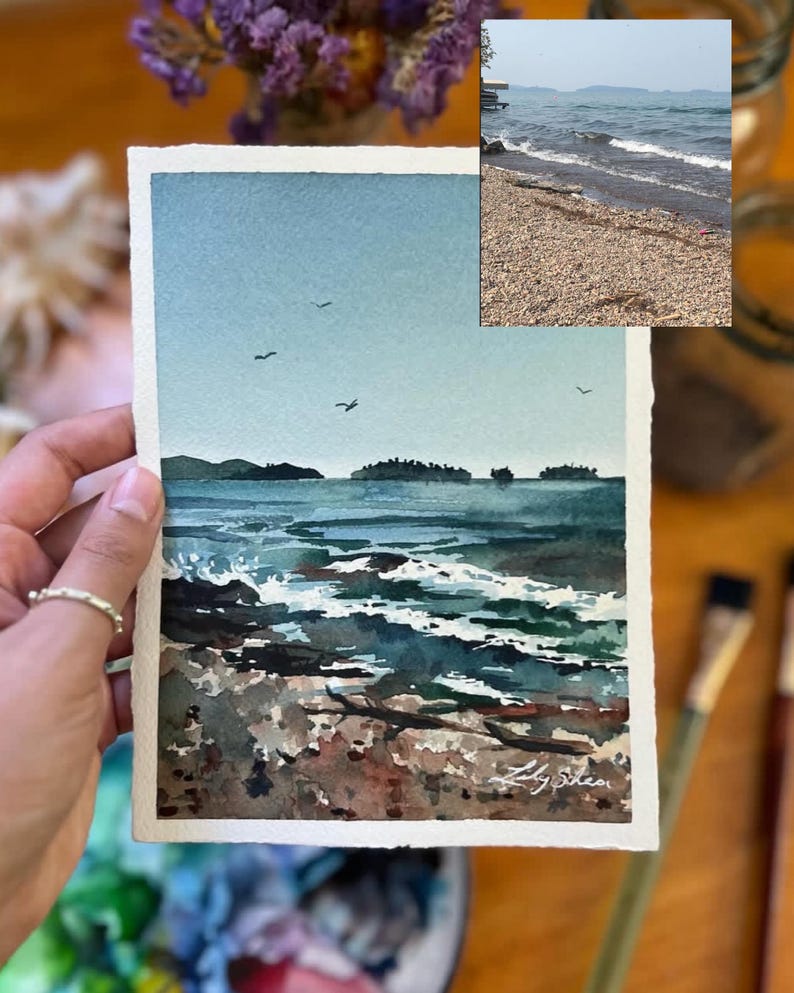 Puede incluir: Pintura acuarela de un paisaje mar&iacute;timo con una costa rocosa, agua azul y algunas aves volando en el cielo. La pintura est&aacute; firmada "Lily Shea" en la esquina inferior derecha.