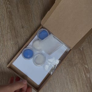 May include: An open cardboard box containing small containers of blue and white paint, a white sponge, and a clear plastic bag. The box is held by hands, and the contents are arranged on a white sheet of paper.