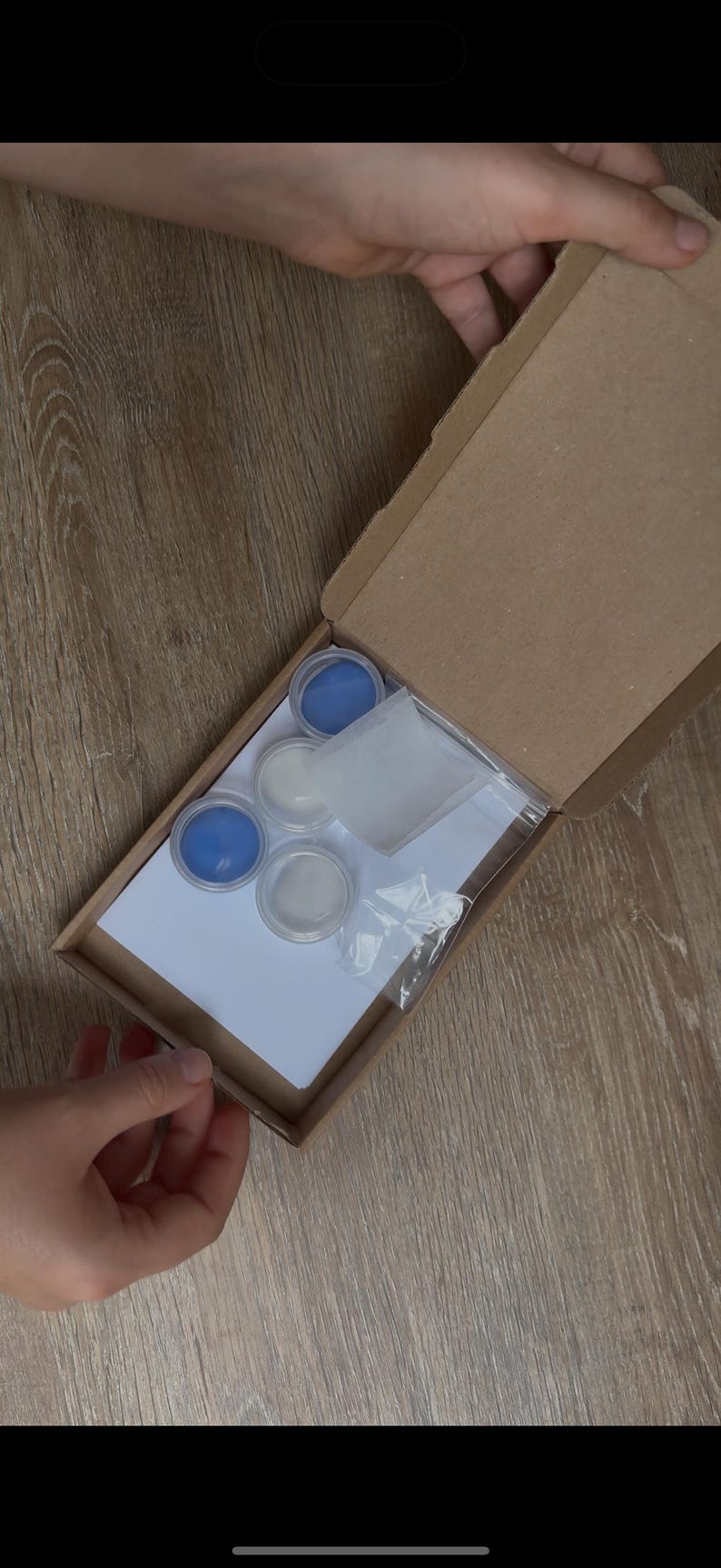 May include: An open cardboard box containing small containers of blue and white paint, a white sponge, and a clear plastic bag. The box is held by hands, and the contents are arranged on a white sheet of paper.