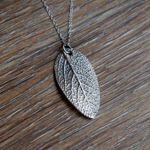 May include: A silver-colored leaf pendant necklace. The pendant is a detailed replica of a leaf, showcasing intricate veining. The necklace chain is delicate and silver, creating an elegant accessory. The pendant is approximately 2 inches long.