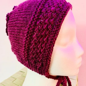 May include: A deep purple knitted hat with a tie at the bottom. The hat has a unique design with a textured pattern.