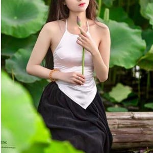 May include: A person wearing a white halter top and a black skirt, holding a lotus bud. The background features large green lily pads. The image has a soft focus and natural lighting, creating a serene atmosphere.