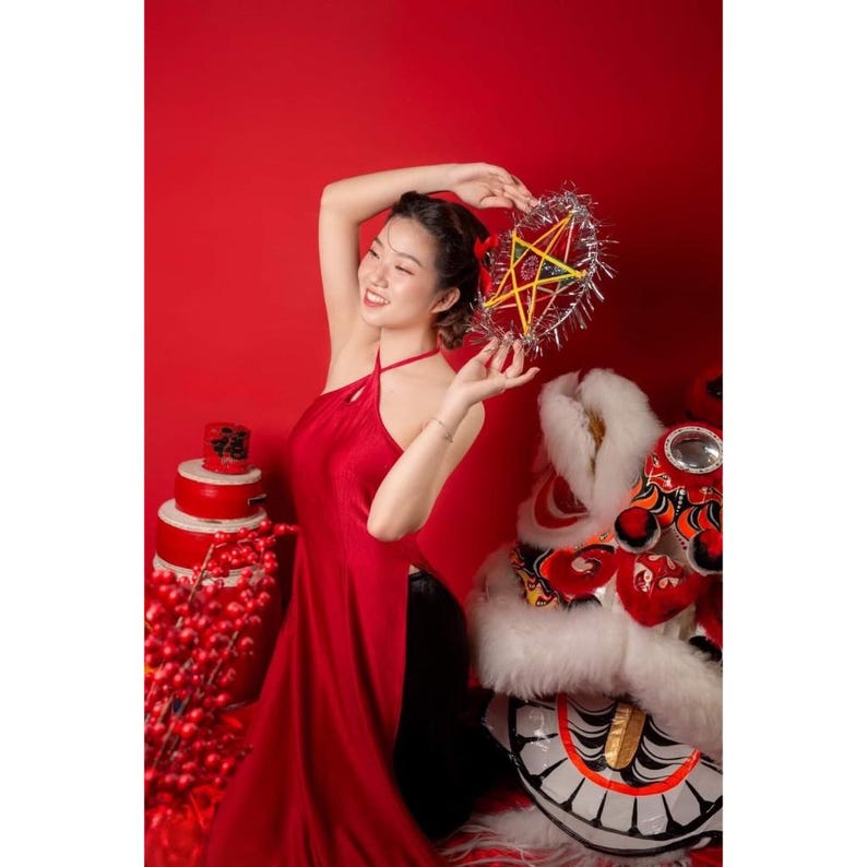 May include: A woman in a red halter dress holds a star-shaped lantern with silver tinsel trim. A red background features a stack of red containers and a lion dance head. The image evokes a festive, cultural theme.
