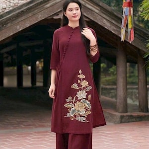 May include: A deep burgundy tunic and wide-leg trousers set. The tunic features a mandarin collar, a side-button closure with a row of small fabric buttons, three-quarter sleeves, and an embroidered floral design in gold and white on the front.