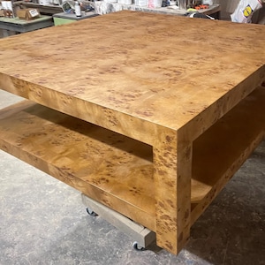 May include: A large square coffee table crafted from light brown wood with a prominent burl grain pattern. The table features a thick flat top surface and a lower shelf supported by four sturdy corner legs, resting on a small wheeled dolly base.