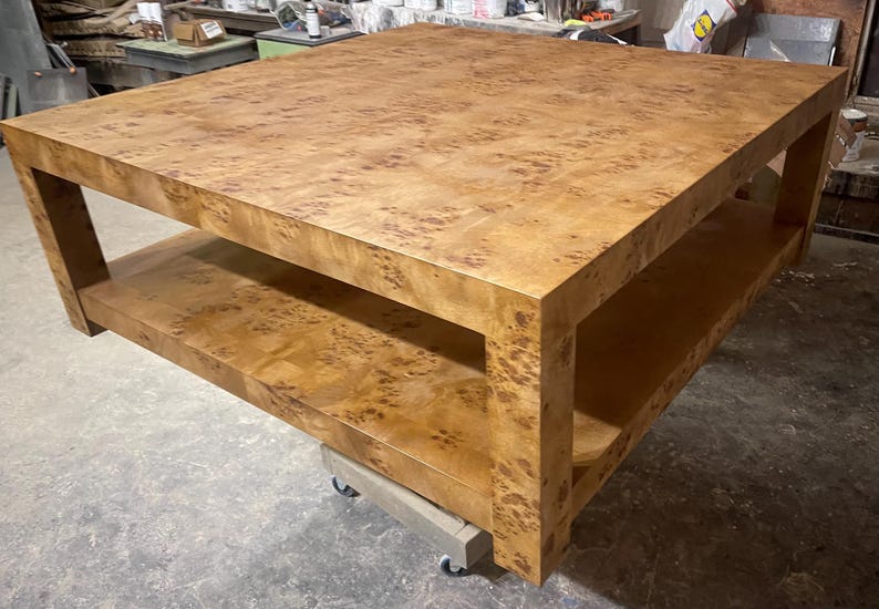 May include: A large square coffee table crafted from light brown wood with a prominent burl grain pattern. The table features a thick flat top surface and a lower shelf supported by four sturdy corner legs, resting on a small wheeled dolly base.