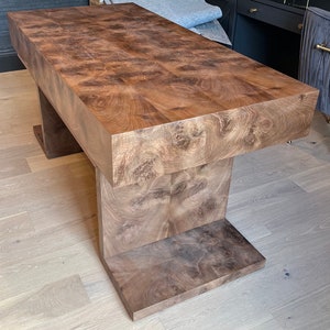 May include: A large, rectangular wooden coffee table with a unique, intricate wood grain pattern. The table has a thick, solid top and a base with two stepped supports.