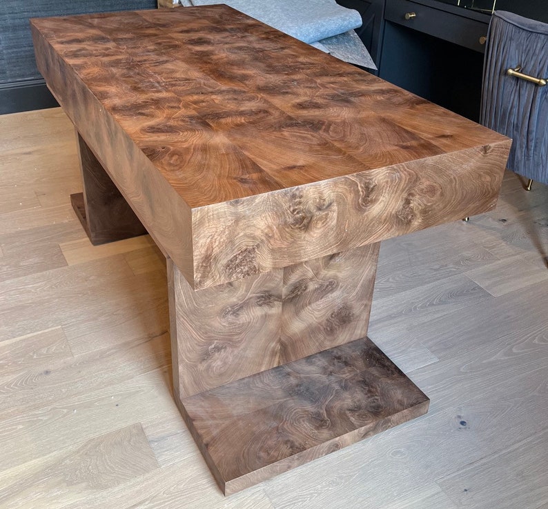 May include: A large, rectangular wooden coffee table with a unique, intricate wood grain pattern. The table has a thick, solid top and a base with two stepped supports.