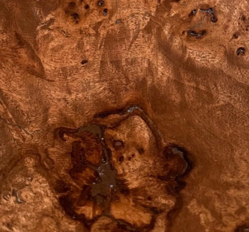 May include: A close-up view of a polished wooden surface featuring a natural burl grain pattern. The wood displays a rich, warm brown color with intricate, swirling dark brown knots and organic textures throughout the entire visible area.