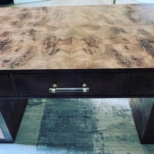 May include: A dark wood desk with three drawers and a unique wood grain pattern on the top. The desk has a modern design with a geometric base and clear acrylic drawer pulls.