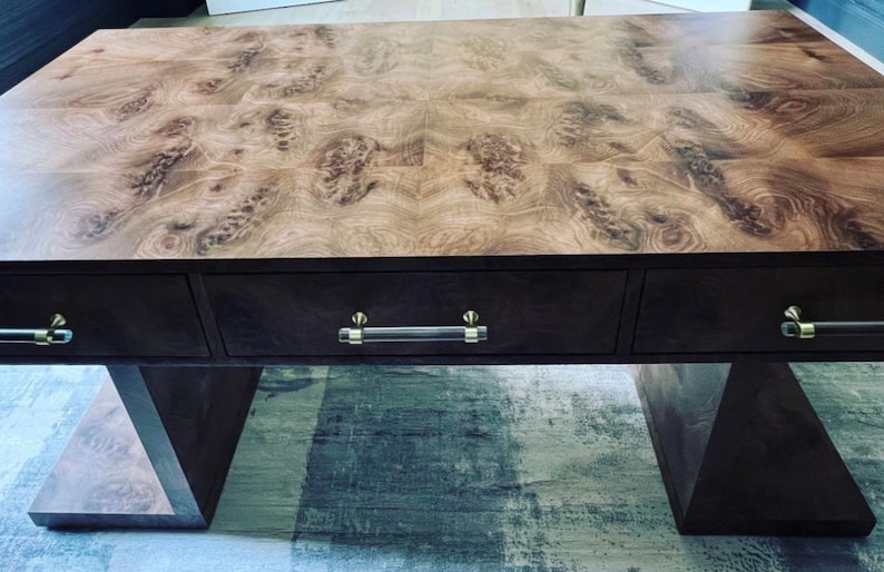 May include: A dark wood desk with three drawers and a unique wood grain pattern on the top. The desk has a modern design with a geometric base and clear acrylic drawer pulls.