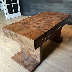 May include: A dark brown wooden desk with three drawers and brass pulls. The desk has a unique, modern design with a wide, flat top and a base that resembles an upside-down 'T'.