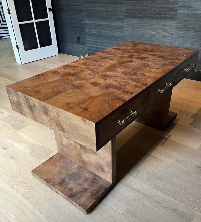 May include: A dark brown wooden desk with three drawers and brass pulls. The desk has a unique, modern design with a wide, flat top and a base that resembles an upside-down 'T'.