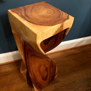 May include: A natural wood side table featuring a square top with visible grain patterns and a twisted, sculptural base. The piece is crafted from a single block of wood, showcasing light and dark brown tones with a smooth, polished finish throughout.