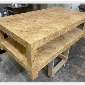 May include: A rectangular coffee table crafted from light-colored burl wood featuring a distinct swirling grain pattern. The table includes a solid flat top surface and a lower shelf supported by four thick, sturdy legs, showcasing a natural finish.