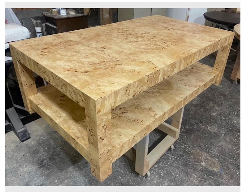 May include: A rectangular coffee table crafted from light-colored burl wood featuring a distinct swirling grain pattern. The table includes a solid flat top surface and a lower shelf supported by four thick, sturdy legs, showcasing a natural finish.