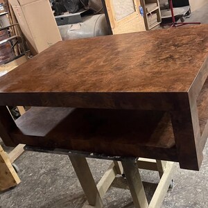 May include: A square coffee table featuring a rich, dark brown burl wood grain finish. The table has a thick, solid top surface and a matching lower shelf, supported by four sturdy, straight legs that connect the top and bottom sections.