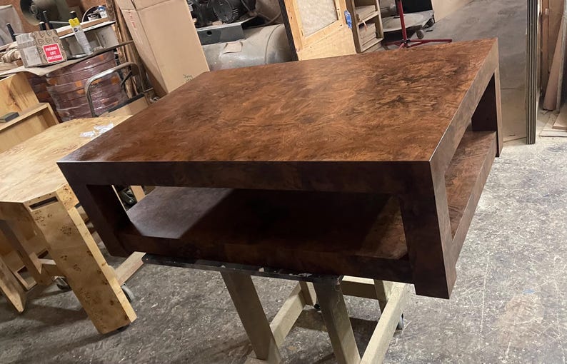 May include: A square coffee table featuring a rich, dark brown burl wood grain finish. The table has a thick, solid top surface and a matching lower shelf, supported by four sturdy, straight legs that connect the top and bottom sections.