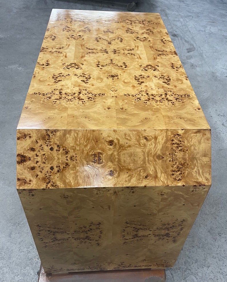 May include: A large, rectangular wooden cabinet with a burl wood veneer. The cabinet has a light brown color with a distinctive pattern of knots and swirls. The cabinet is made of solid wood and has a smooth, polished finish.