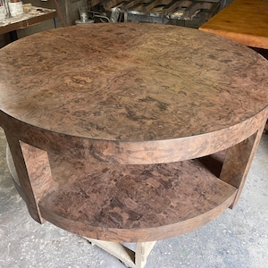 May include: A round coffee table featuring a rich brown burl wood grain pattern across the circular top and the lower shelf. The table is supported by thick, curved vertical legs that connect the top surface to the matching circular base shelf.