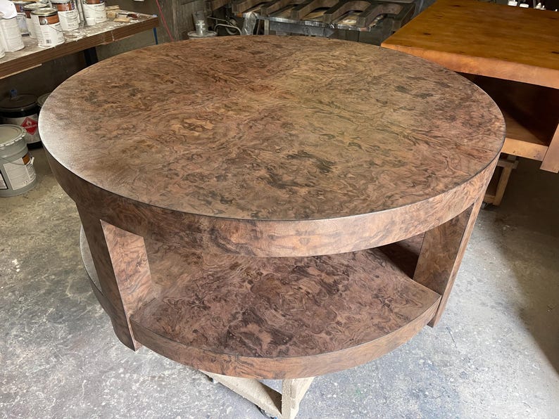 May include: A round coffee table featuring a rich brown burl wood grain pattern across the circular top and the lower shelf. The table is supported by thick, curved vertical legs that connect the top surface to the matching circular base shelf.