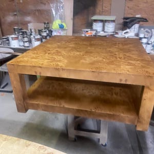 May include: A square coffee table crafted from light brown burl wood with a distinct swirling grain pattern. The table features a thick, flat top surface and a lower open shelf supported by four sturdy, straight legs, resting on a mobile base.