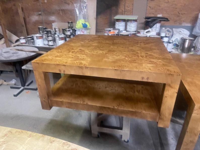 May include: A square coffee table crafted from light brown burl wood with a distinct swirling grain pattern. The table features a thick, flat top surface and a lower open shelf supported by four sturdy, straight legs, resting on a mobile base.