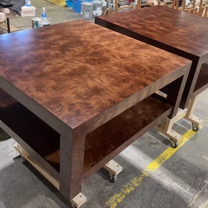 May include: Two square coffee tables featuring a rich, dark brown burl wood grain pattern. Each table has a flat top surface and a lower shelf supported by four thick, sturdy legs, providing a clean, geometric silhouette for the furniture.