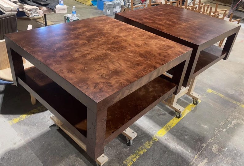 May include: Two square coffee tables featuring a rich, dark brown burl wood grain pattern. Each table has a flat top surface and a lower shelf supported by four thick, sturdy legs, providing a clean, geometric silhouette for the furniture.