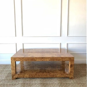May include: A square coffee table featuring a polished burl wood veneer with a distinct swirling grain pattern. The table has a thick, block-style frame with four sturdy legs and a lower shelf that provides an additional flat surface for storage.
