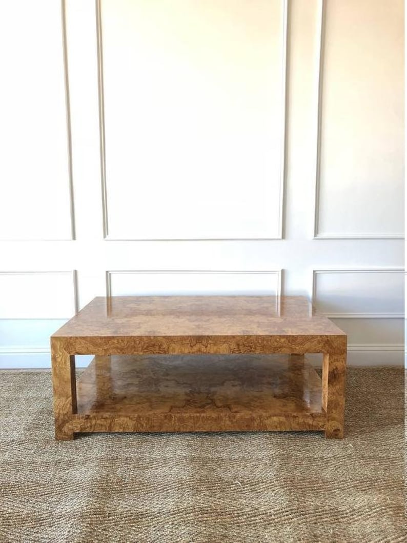 May include: A square coffee table featuring a polished burl wood veneer with a distinct swirling grain pattern. The table has a thick, block-style frame with four sturdy legs and a lower shelf that provides an additional flat surface for storage.
