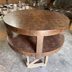 May include: A round, two-tiered table featuring a rich, dark brown burl wood veneer finish. The table has a circular top surface and a matching circular lower shelf, both supported by thick, vertical wooden legs connecting the two levels.