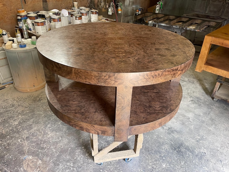 May include: A round, two-tiered table featuring a rich, dark brown burl wood veneer finish. The table has a circular top surface and a matching circular lower shelf, both supported by thick, vertical wooden legs connecting the two levels.