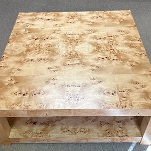 May include: A square coffee table constructed from light-colored wood featuring a prominent burl grain pattern. The table has a smooth, flat top surface and a lower shelf, both showcasing the natural, swirling, and spotted wood grain texture.
