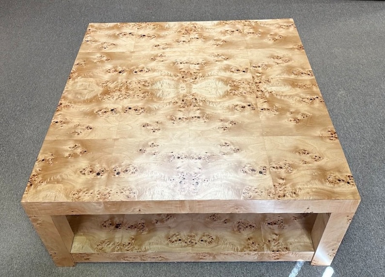 May include: A square coffee table constructed from light-colored wood featuring a prominent burl grain pattern. The table has a smooth, flat top surface and a lower shelf, both showcasing the natural, swirling, and spotted wood grain texture.