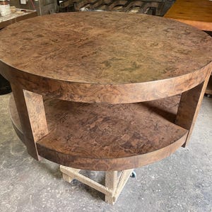 May include: A round, two-tiered table crafted from brown wood with a prominent burl grain pattern. The table features a circular top surface and a matching circular lower shelf, both supported by thick, vertical wooden legs in a matching finish.